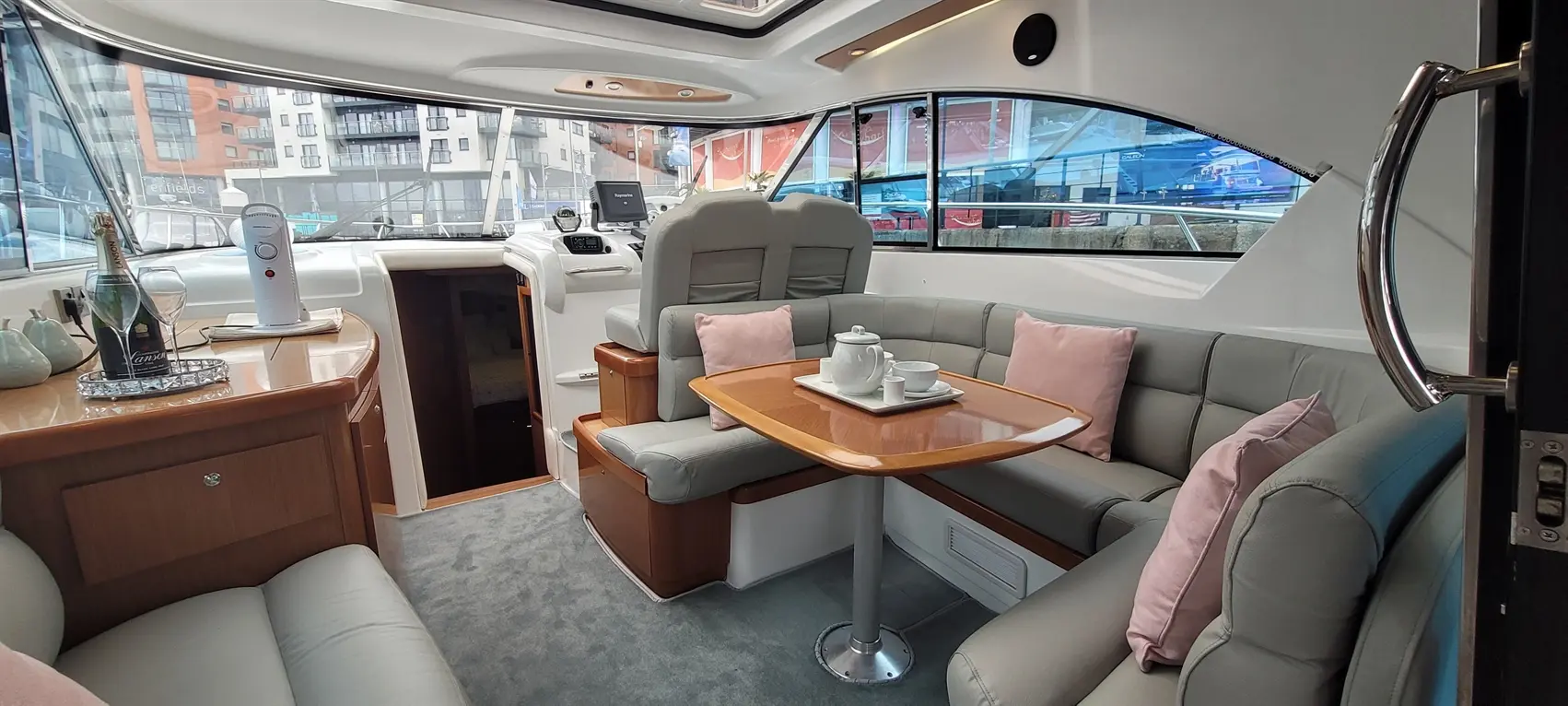Luxurious boat interior featuring a seating area, dining table, champagne, and views of the marina through large windows.